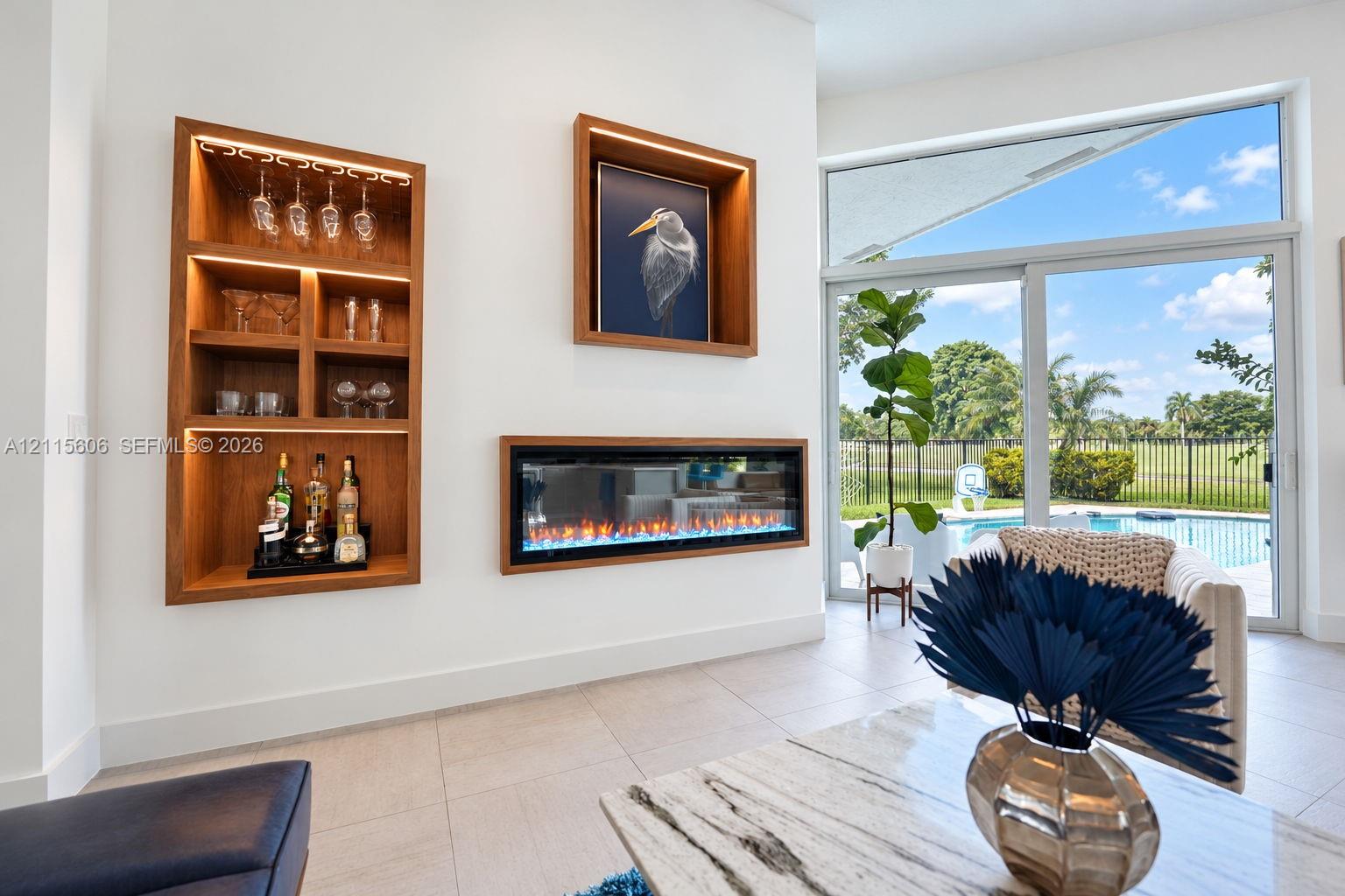 Built-in illuminated wood shelving, recessed linear electric fireplace, large-format tile flooring, and floor-to-ceiling sliding glass doors with outdoor pool exposure