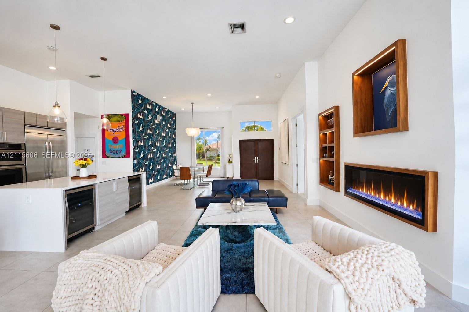 Open-concept interior featuring large-format tile flooring, a wall-mounted linear electric fireplace with wood trim, built-in recessed shelving, a center island with integrated wine refrigeration, and a patterned accent wall