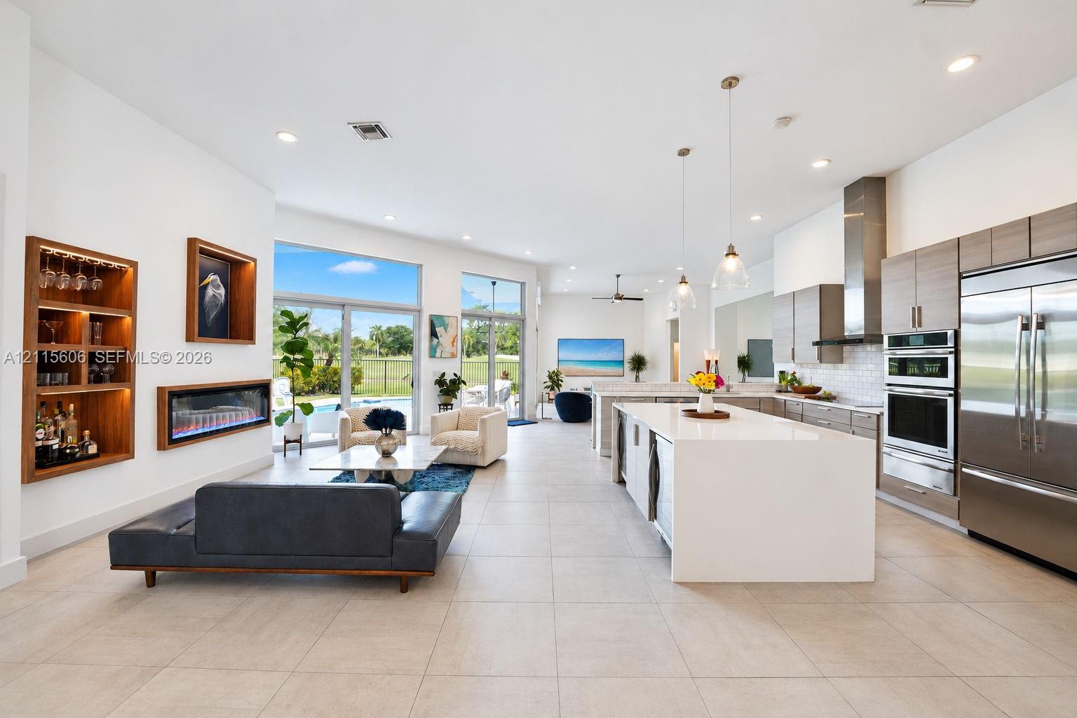 Open-concept living space featuring a central kitchen island with waterfall-edge stone countertop, built-in recessed shelving, integrated linear fireplace, and large glass sliding doors