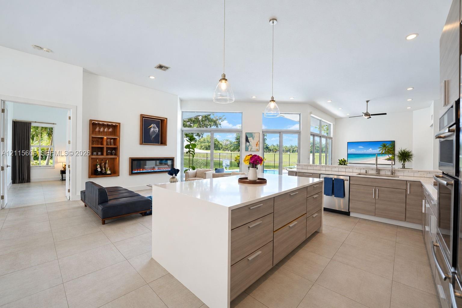Open-concept kitchen featuring an oversized island with quartz countertops, wood-finish cabinetry, built-in stainless steel double ovens, recessed lighting, and a wall-mounted electric fireplace