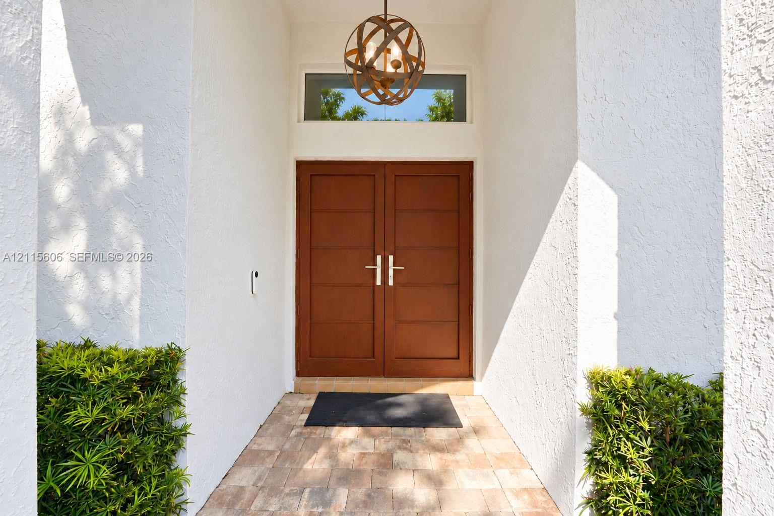 Entryway featuring double wood-finish doors with horizontal paneling, a circular metal orb pendant light fixture, a rectangular transom window, and stone paver flooring