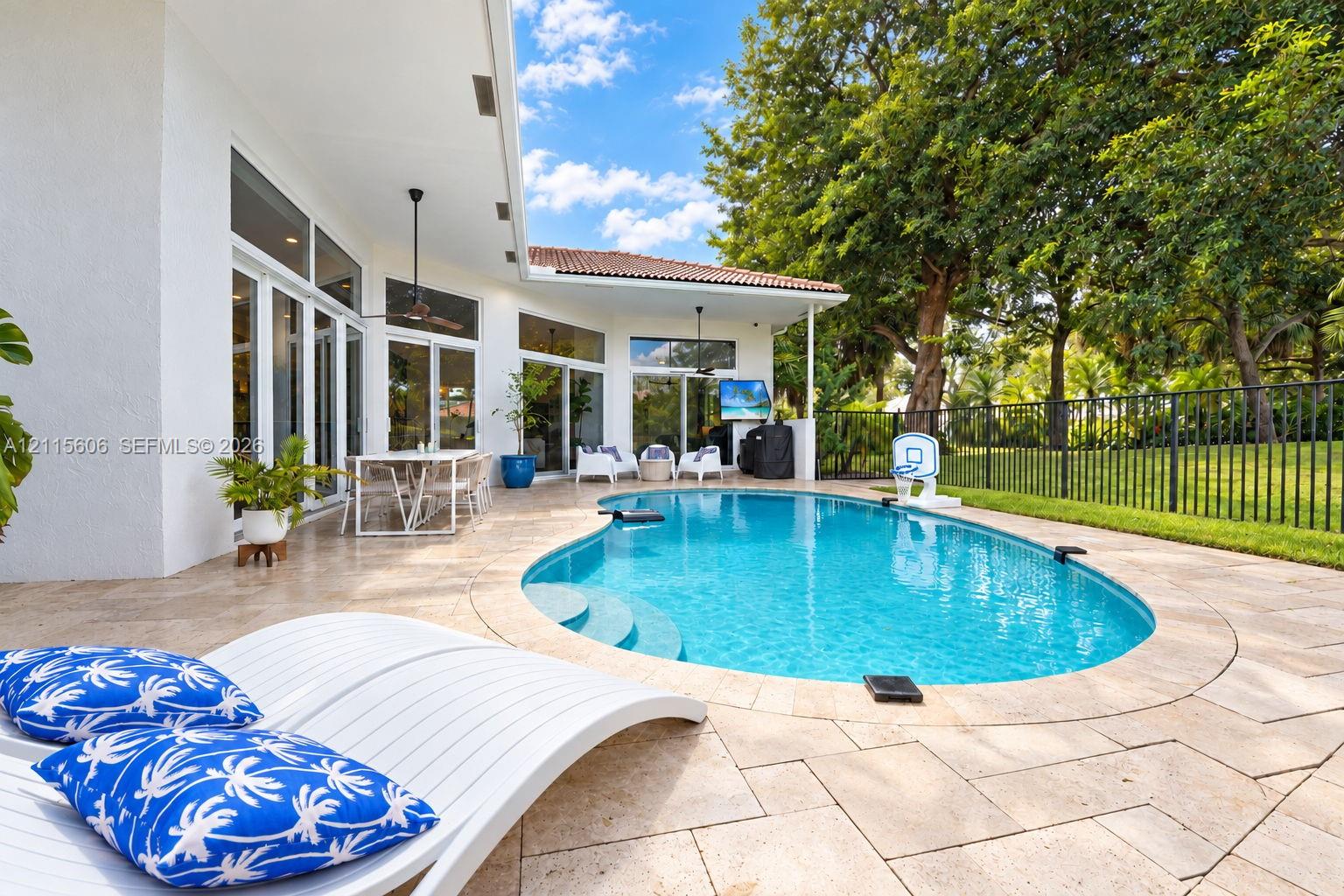 Outdoor patio featuring natural stone pavers, a custom-shaped swimming pool with integrated steps, a covered lanai with ceiling fans, and black metal perimeter fencing