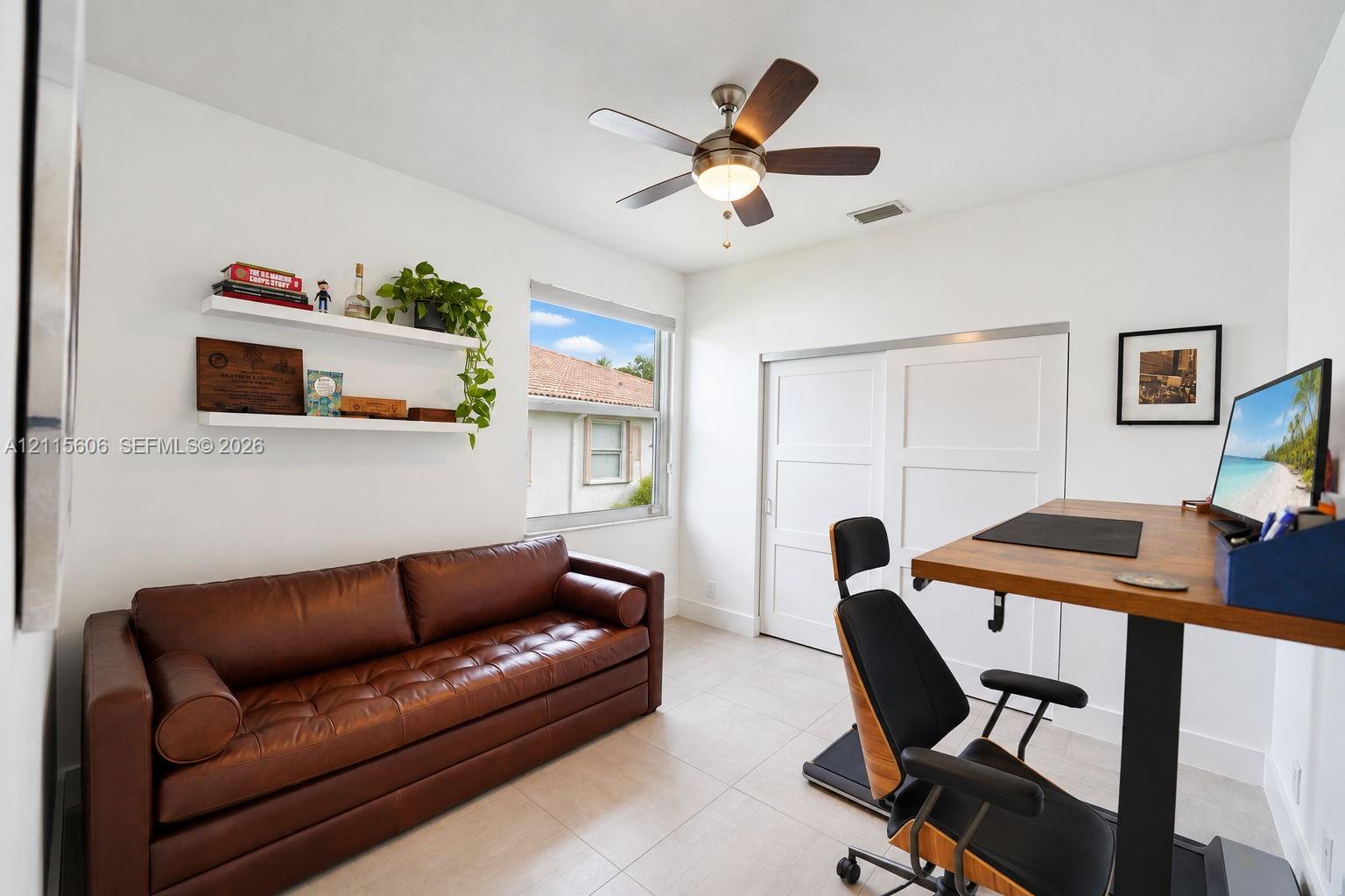 Neutral-toned tile flooring, white sliding closet doors, multi-blade ceiling fan with integrated lighting, and two wall-mounted floating shelves