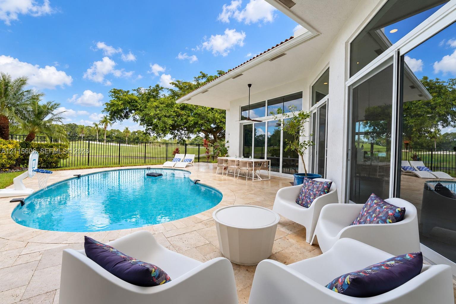 Covered patio with travertine tile flooring, inground swimming pool, floor-to-ceiling glass doors, and black metal perimeter fencing
