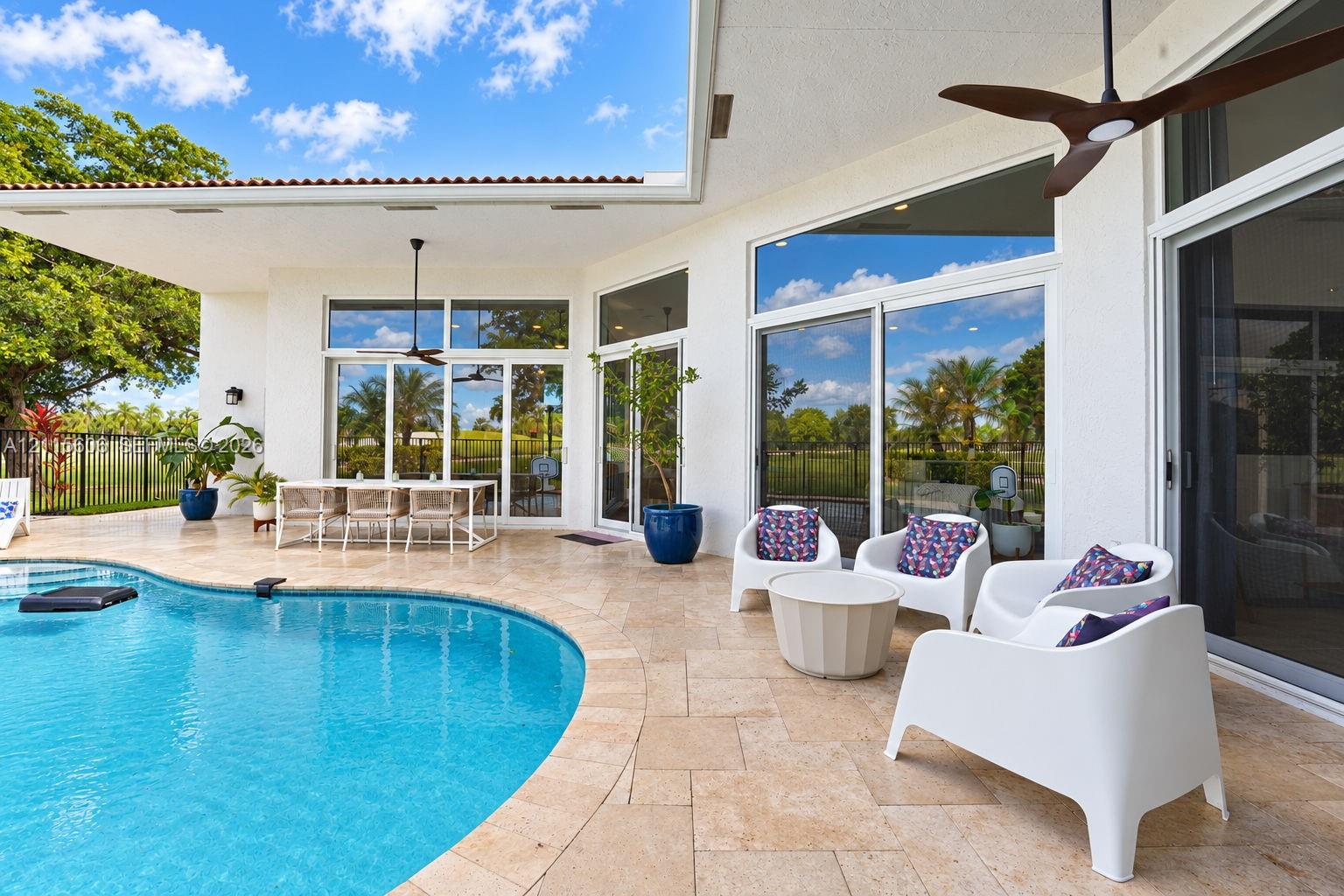 Covered patio featuring stone-tile flooring, ceiling fans, oversized glass sliding doors, and a curved swimming pool