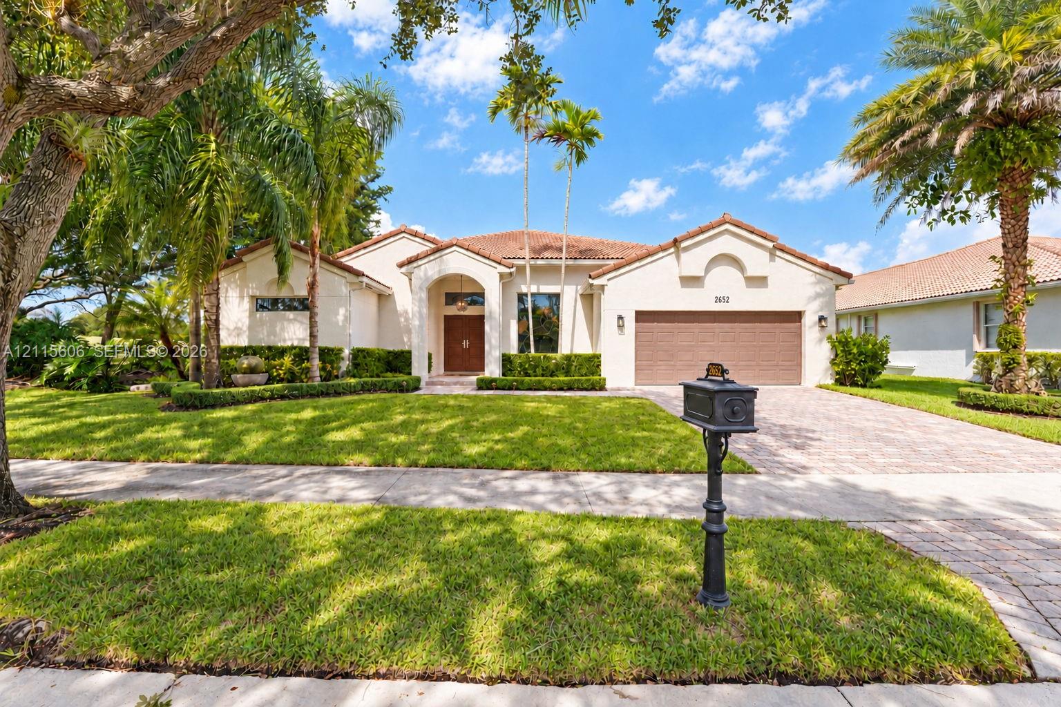 Stucco exterior featuring a clay tile roof, arched entryway, paved driveway, and mature landscaping