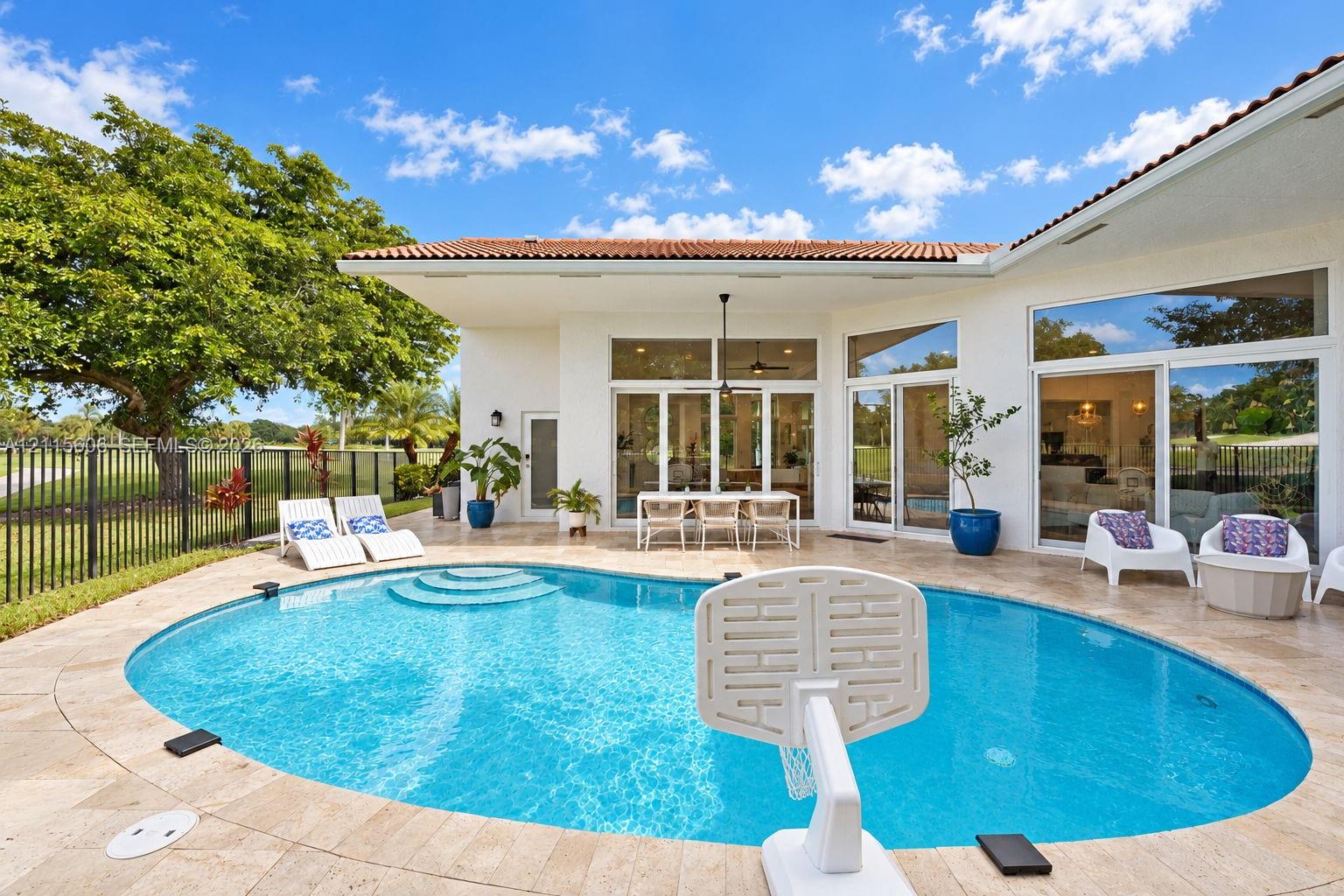 Outdoor patio featuring a travertine stone deck, a curved swimming pool with integrated steps, multiple floor-to-ceiling glass sliding doors, and a covered lanai