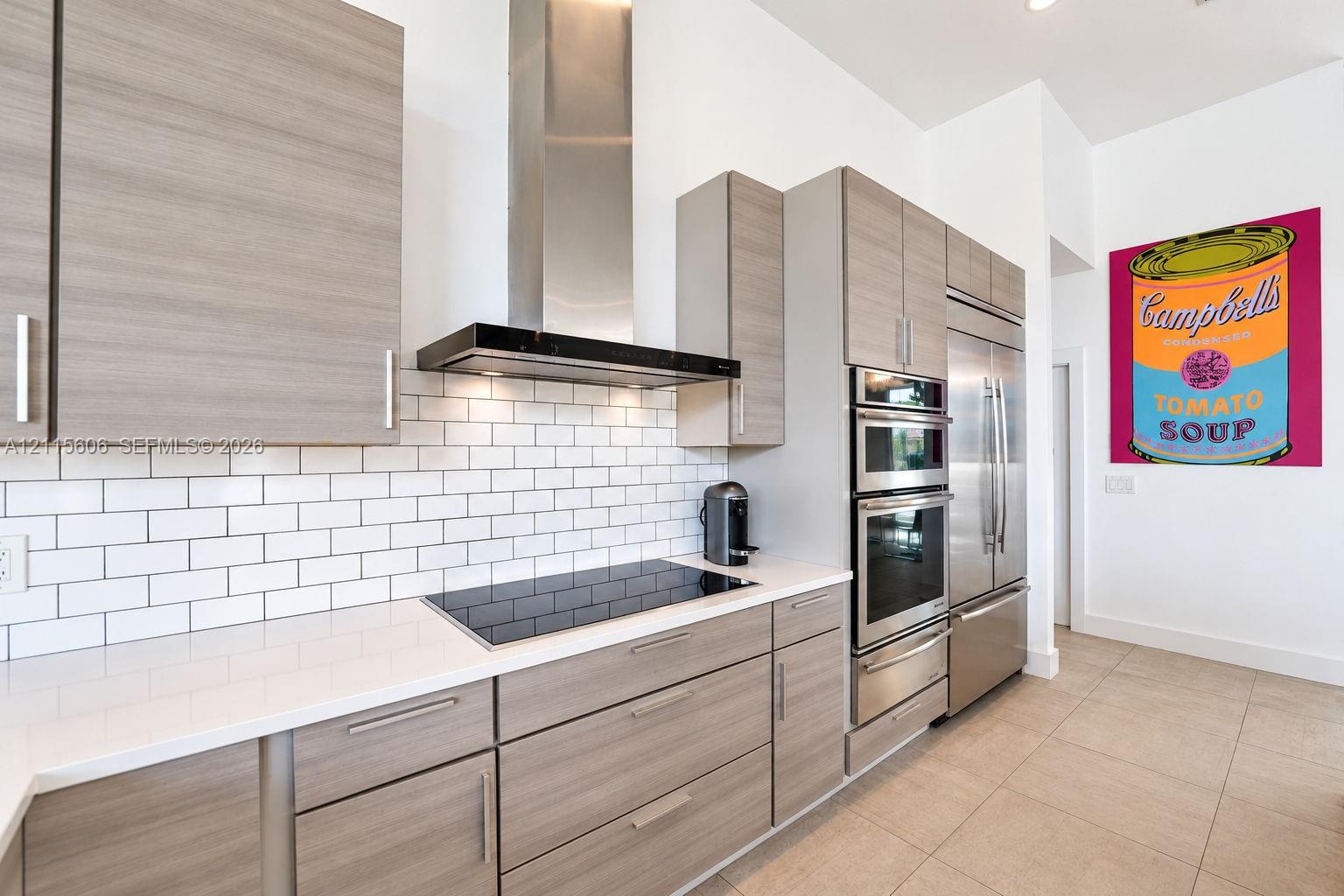 Kitchen featuring light wood-finish cabinetry, white solid-surface countertops, white subway tile backsplash, stainless steel built-in appliances, and a modern chimney-style range hood