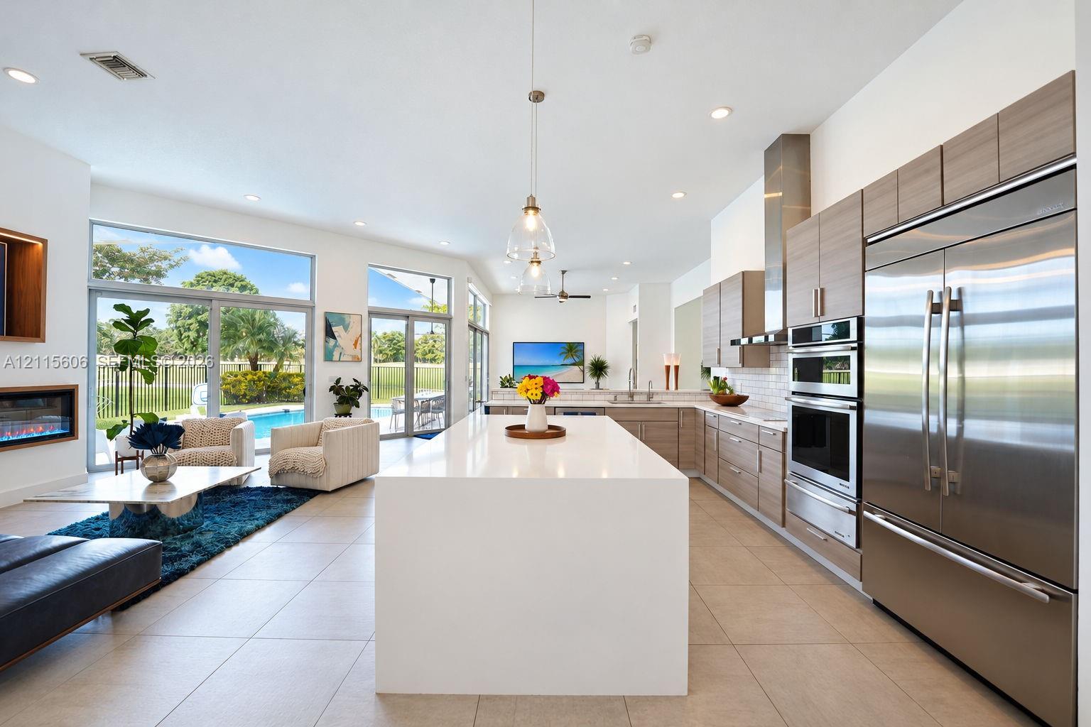 Open-concept interior featuring large center island with solid-surface countertop, linear wood-finish cabinetry, stainless steel built-in appliances, recessed lighting, and expansive sliding glass doors