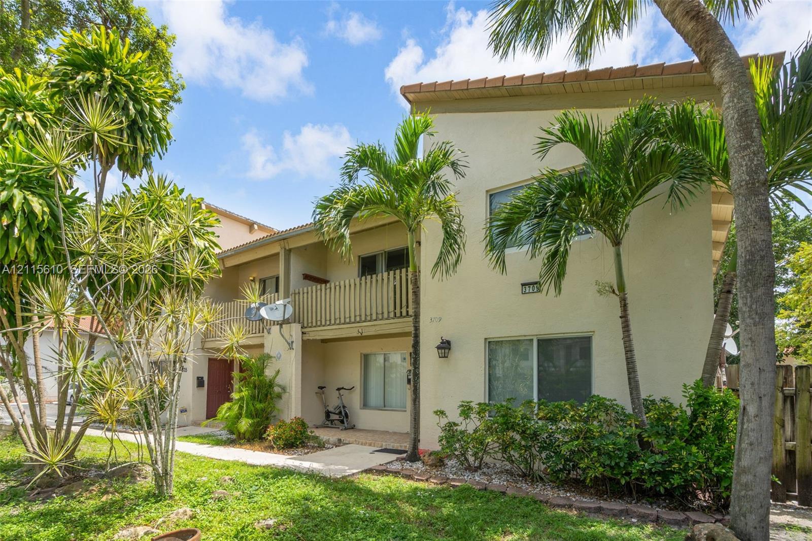 Multi-story exterior featuring light-toned stucco siding, a second-level railed balcony, and mature palm landscaping