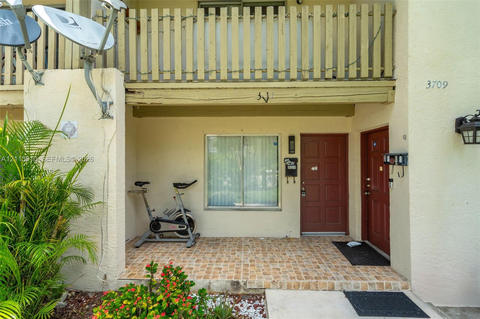Covered entryway featuring a tiled porch, two solid-panel entry doors, a multi-pane slider window, and an overhead balcony with a wooden balustrade