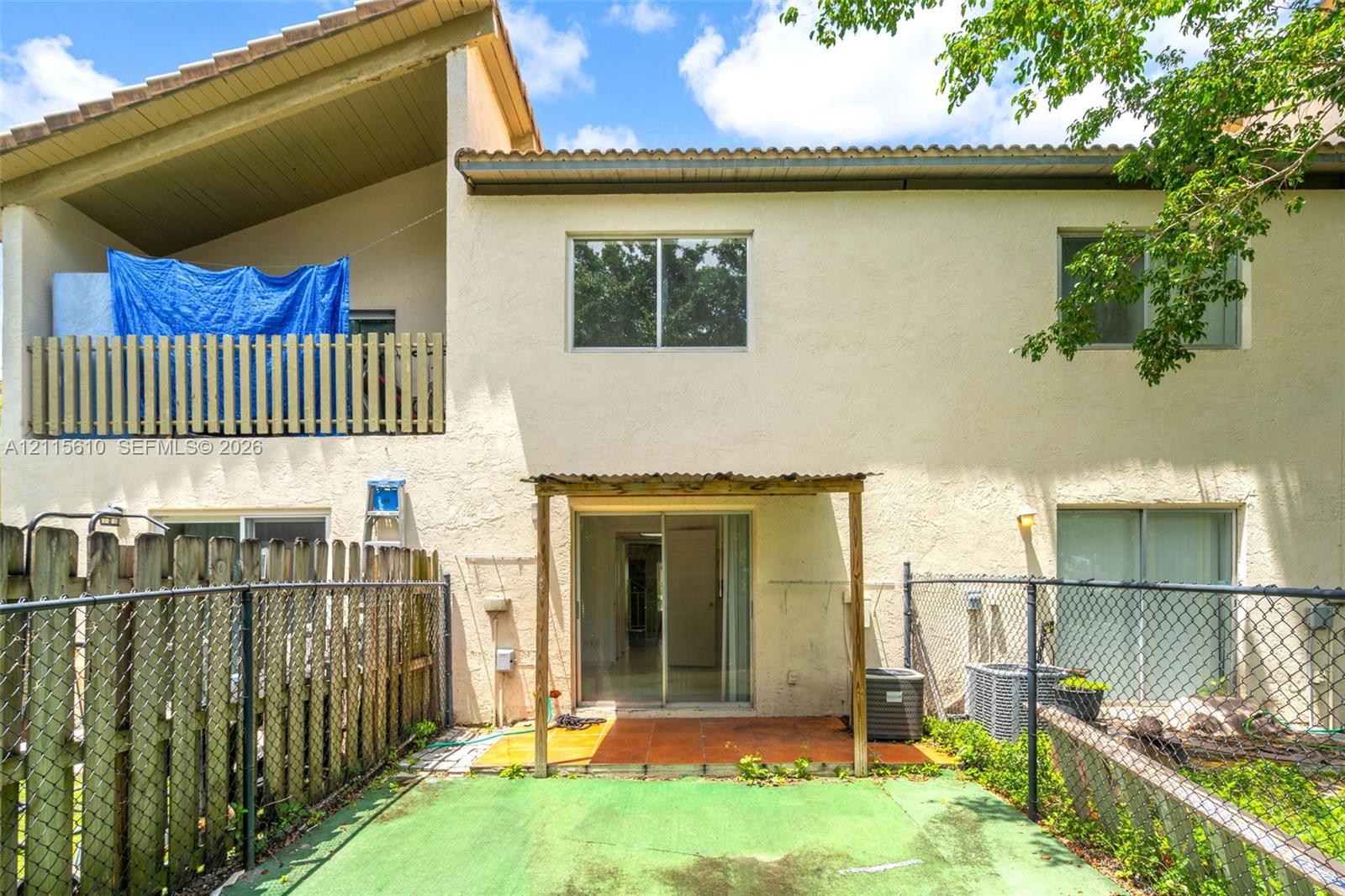 Stucco exterior featuring a covered upper-level balcony with wood-slat railing, a ground-level covered patio with tile flooring, and a fenced yard