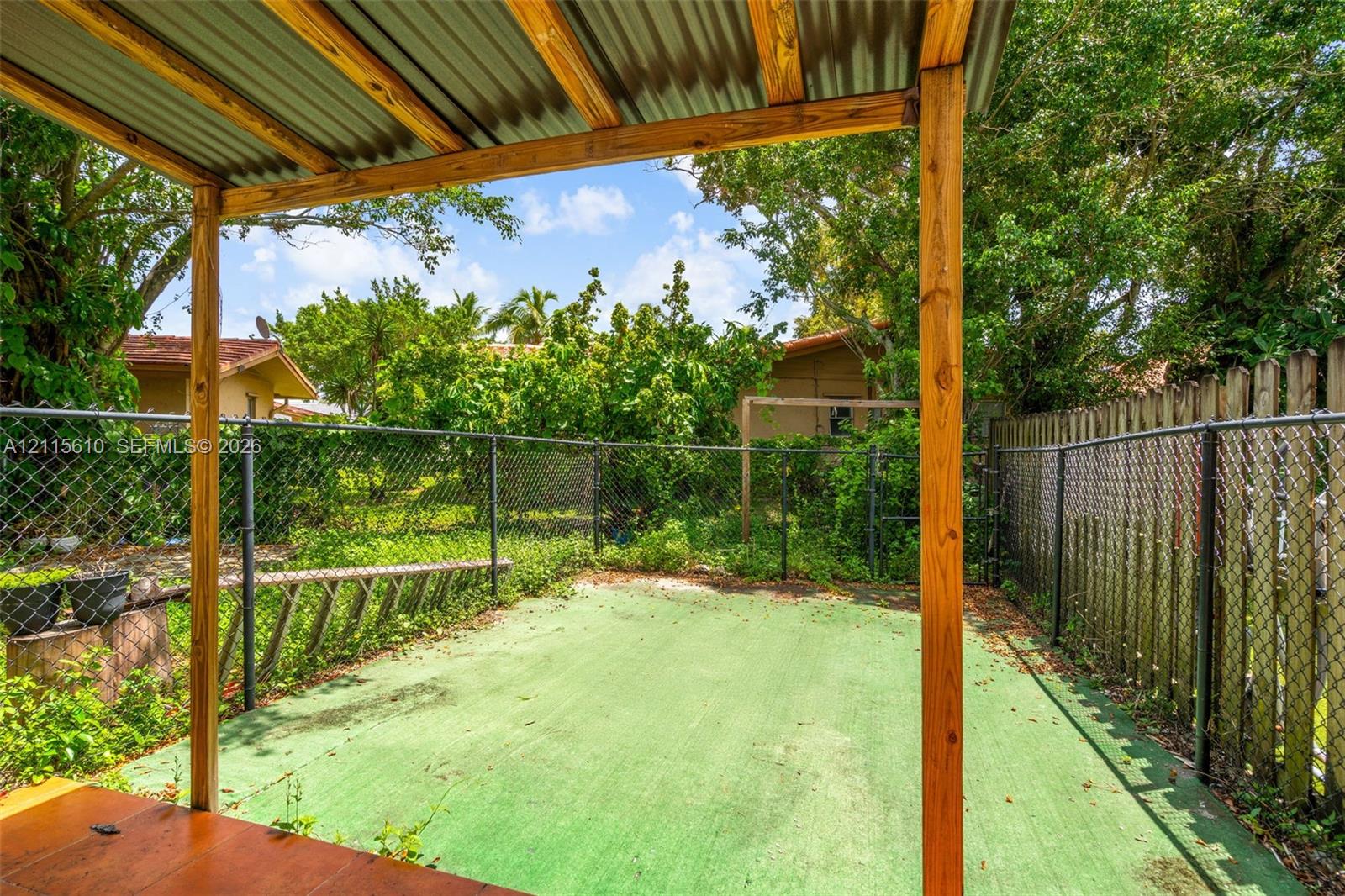 Covered outdoor patio area with a wood-frame structure, metal roofing, green-surfaced concrete flooring, and chain-link fencing