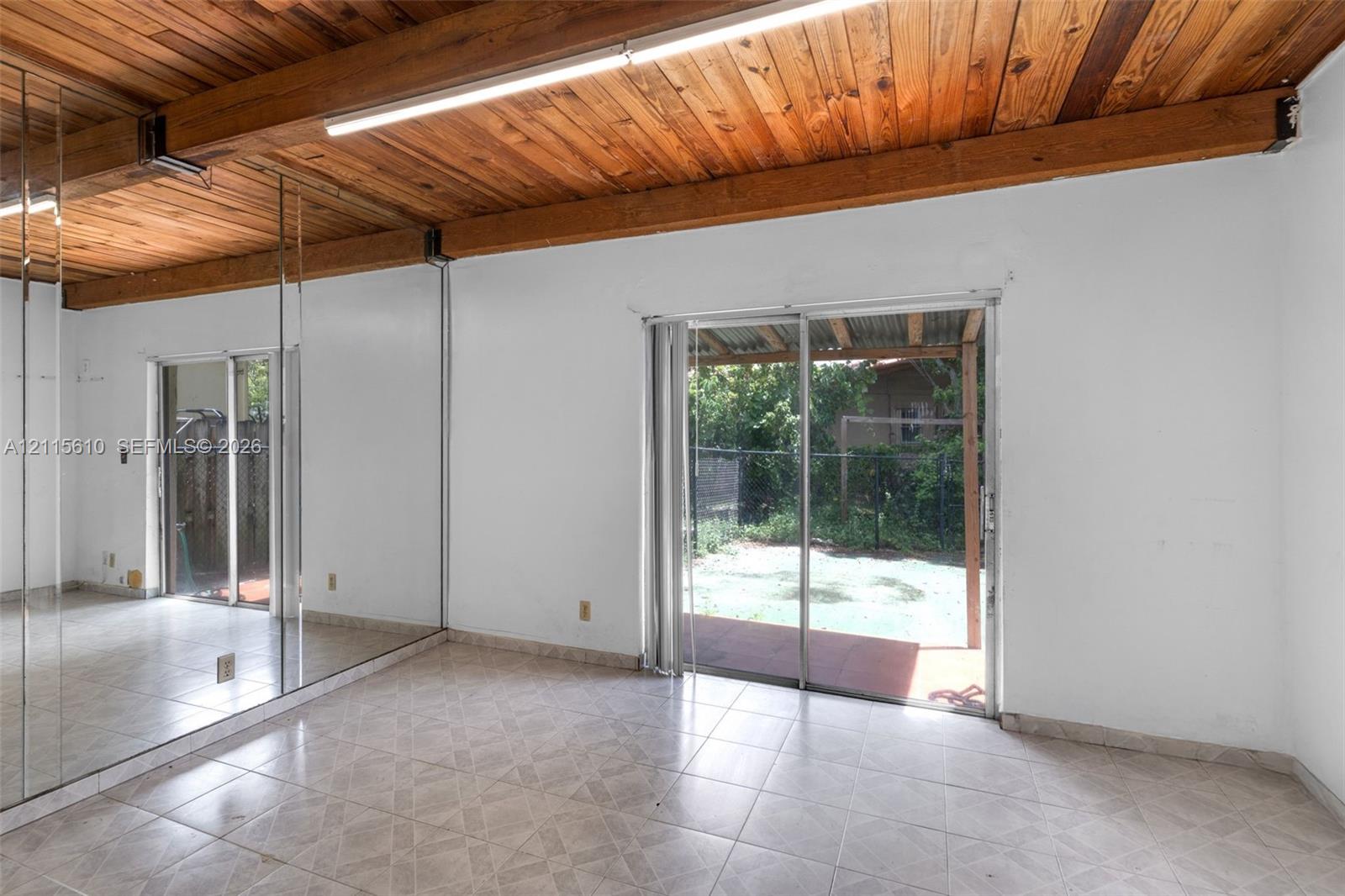 Exposed wood-plank ceiling with structural beams, floor-to-ceiling mirrored walls, ceramic tile flooring, and sliding glass doors leading to a covered patio