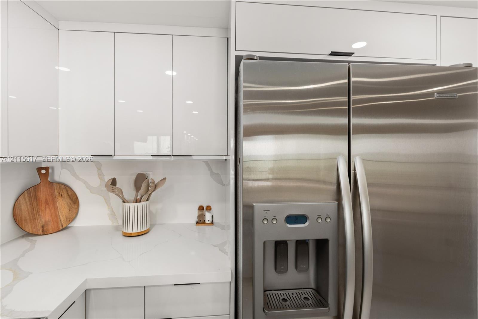 Kitchen featuring high-gloss white cabinetry, white stone-finish countertops with grey veining, full-height stone-finish backsplash, and stainless steel refrigerator with integrated water and ice dispenser
