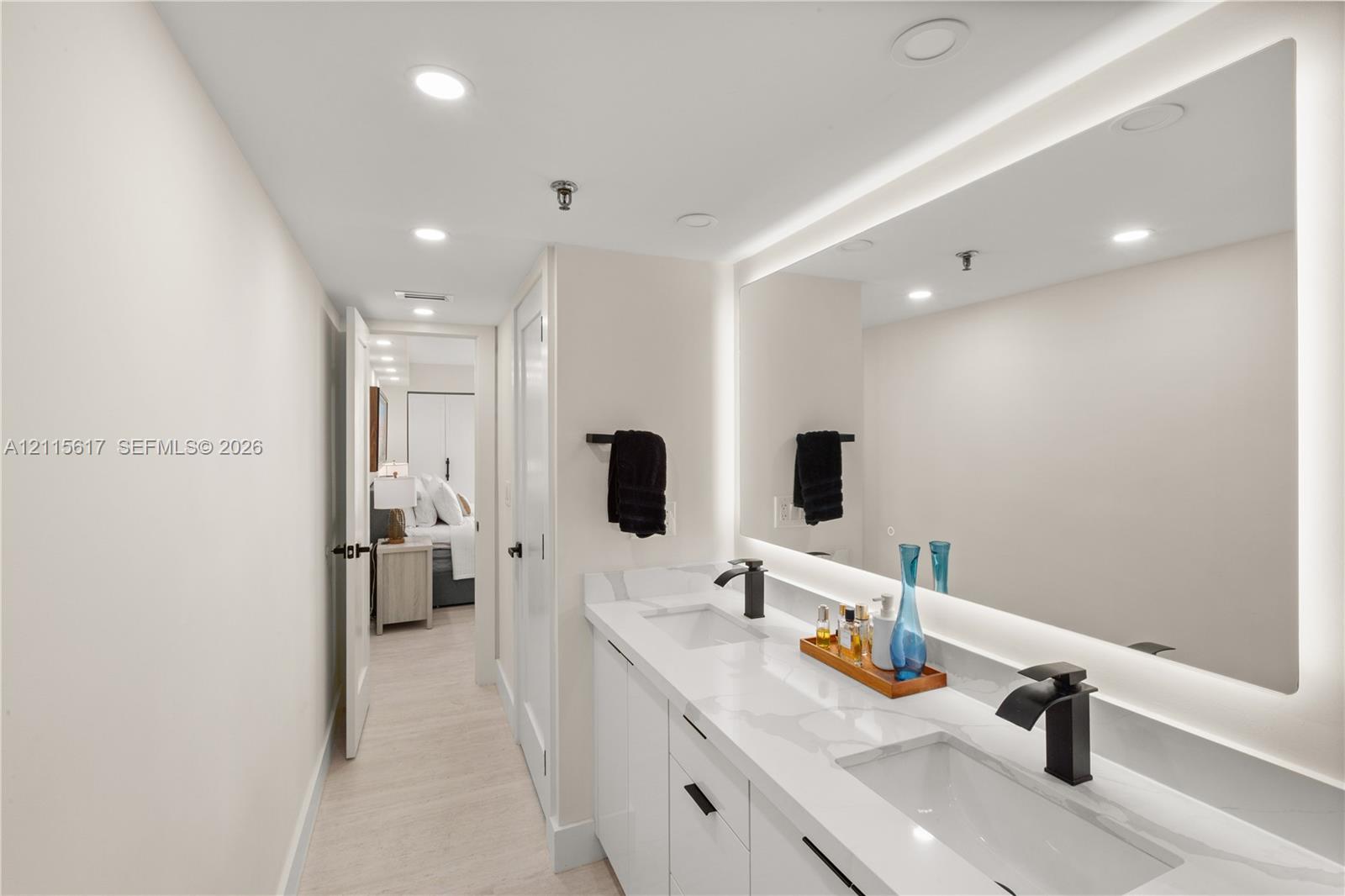 Double vanity featuring a light-toned countertop, integrated rectangular sinks, matte black widespread faucets, and a large backlit mirror