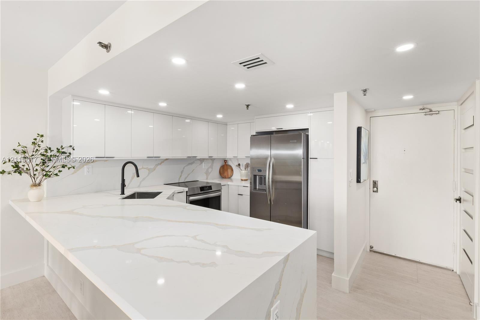 Kitchen featuring white high-gloss cabinetry, stone-finish countertops with waterfall edge, stainless steel refrigerator, and matte black faucet