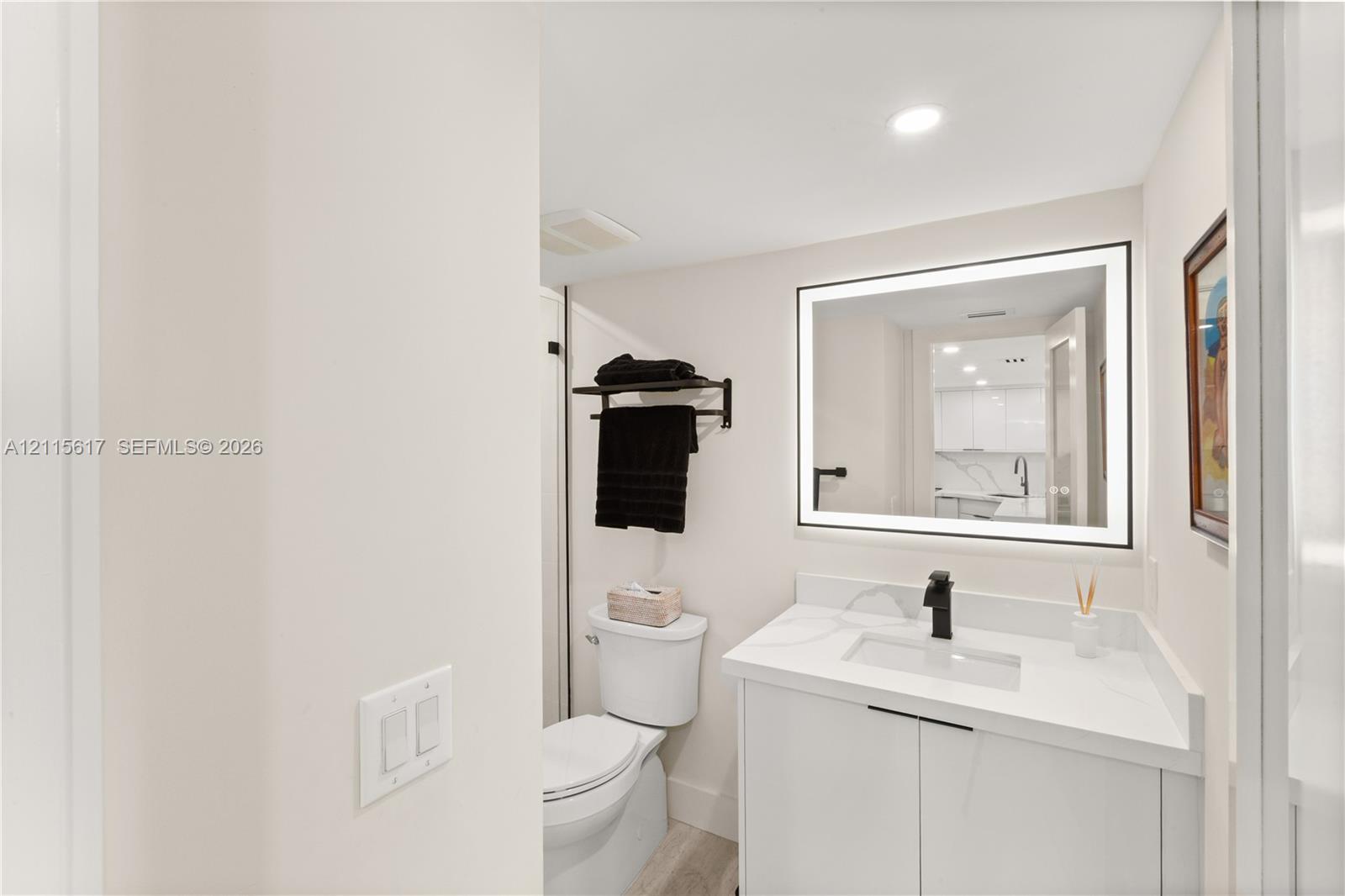 Bathroom featuring a vanity with a white countertop and rectangular integrated sink, matte black faucet, wall-mounted backlit mirror, and two-piece toilet