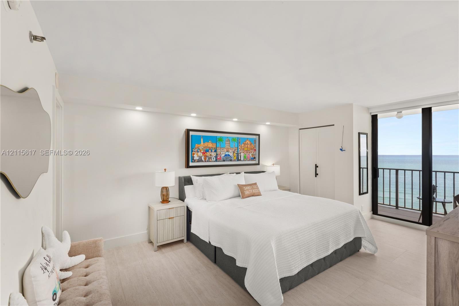 Neutral-toned interior space featuring light-colored flooring, recessed ceiling lighting, a built-in closet with matte black hardware, and floor-to-ceiling glass sliding doors opening to a balcony with an ocean backdrop