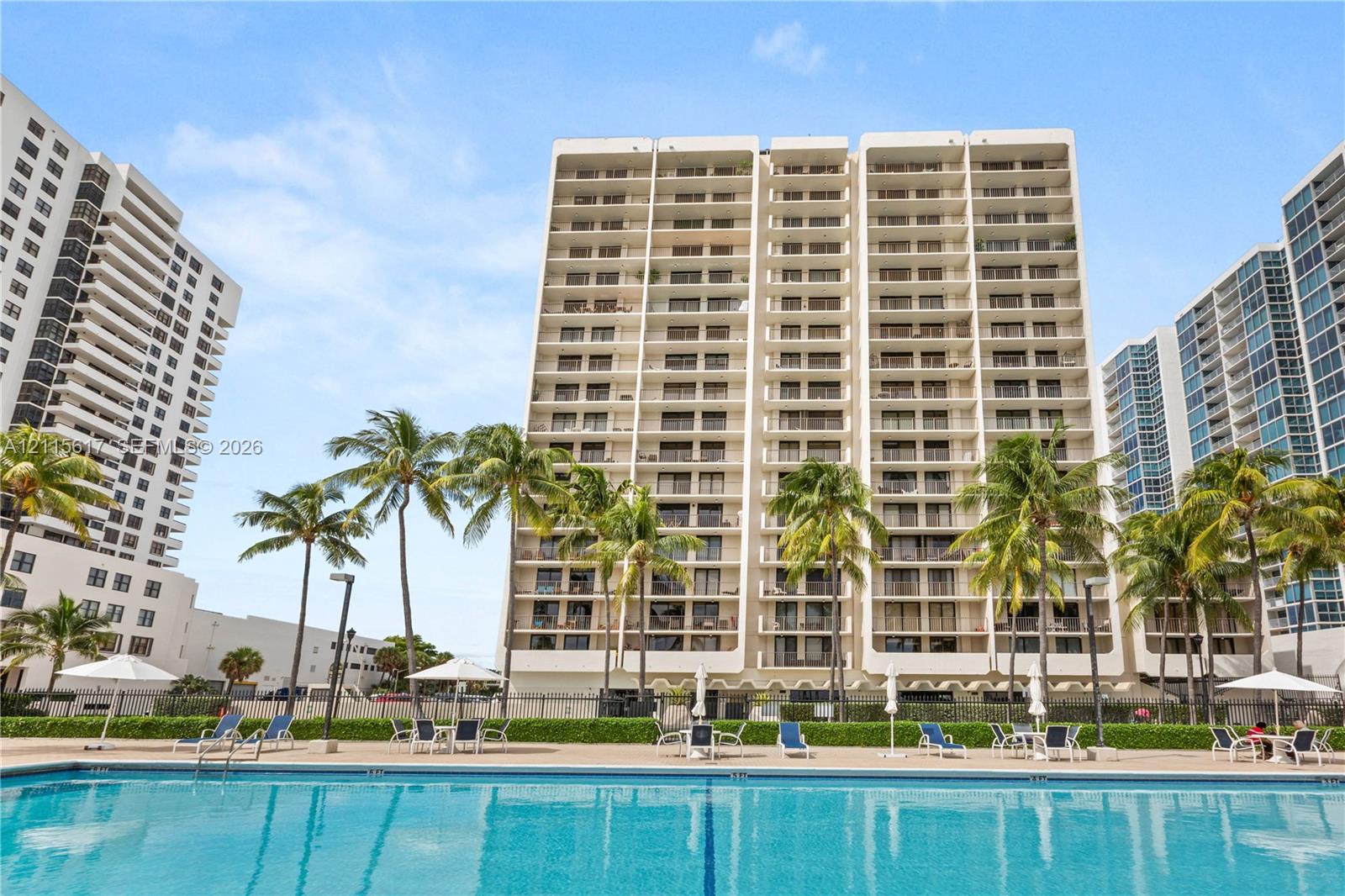 Multi-story high-rise building featuring private balconies, an expansive ground-level swimming pool, and mature palm trees