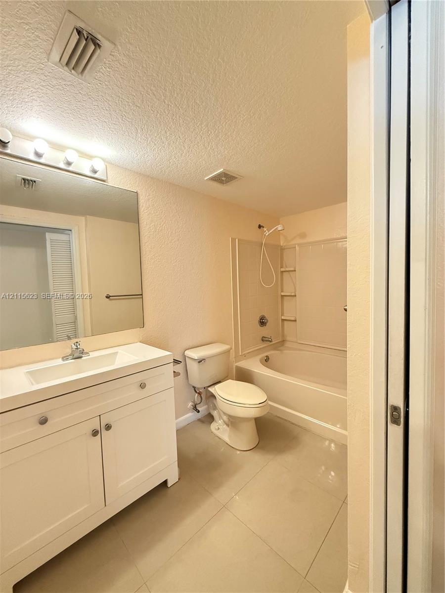 White vanity featuring a single integrated sink, large-format tile flooring, oversized wall mirror with multi-bulb light fixture, and a built-in tub with integrated shelving