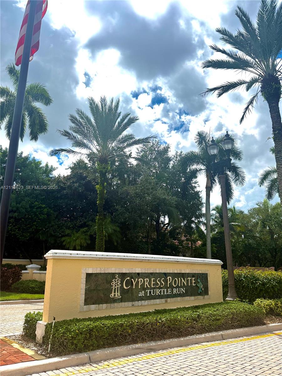 Monument entrance sign for Cypress Pointe at Turtle Run, featuring stucco masonry, stone inlay detailing, manicured hedges, mature palm trees, and an ornate street lamp
