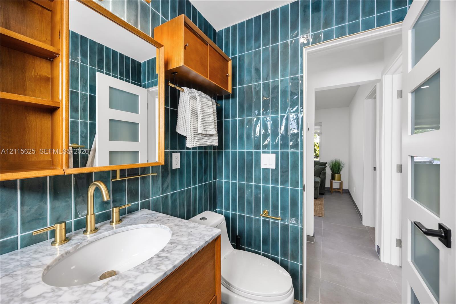 Teal vertical ceramic wall tile, stone countertop with an integrated oval basin, brass-finish widespread faucet, wooden cabinetry featuring built-in shelving, and frosted-glass panel door