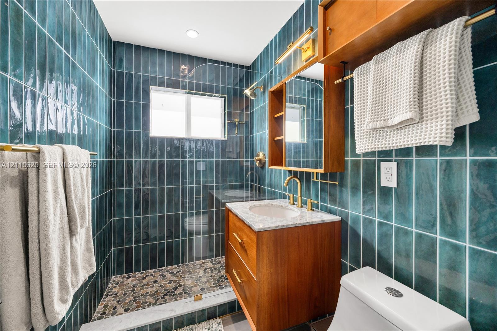 Bathroom featuring floor-to-ceiling vertical teal tiling, stone-finish walk-in shower flooring, built-in wooden cabinetry with a stone countertop, and brass-tone fixtures