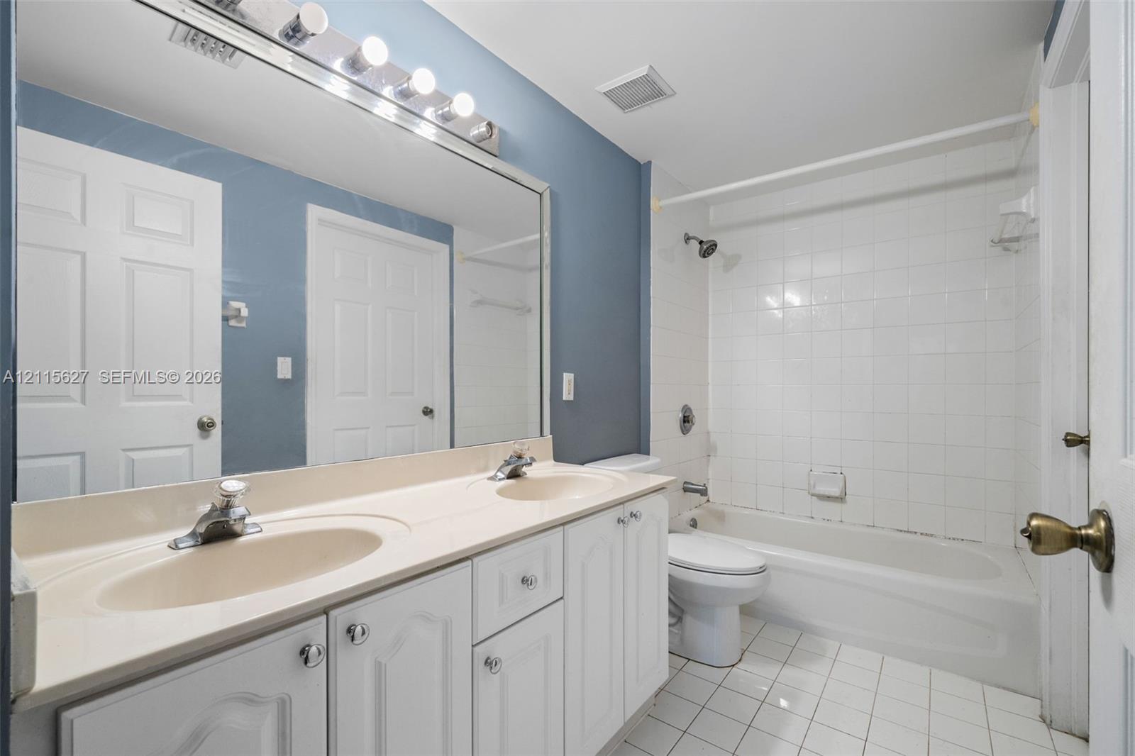 Bathroom featuring a dual-sink vanity with a solid-surface countertop, full-width vanity mirror, tile-surround bathtub, and ceramic tile flooring