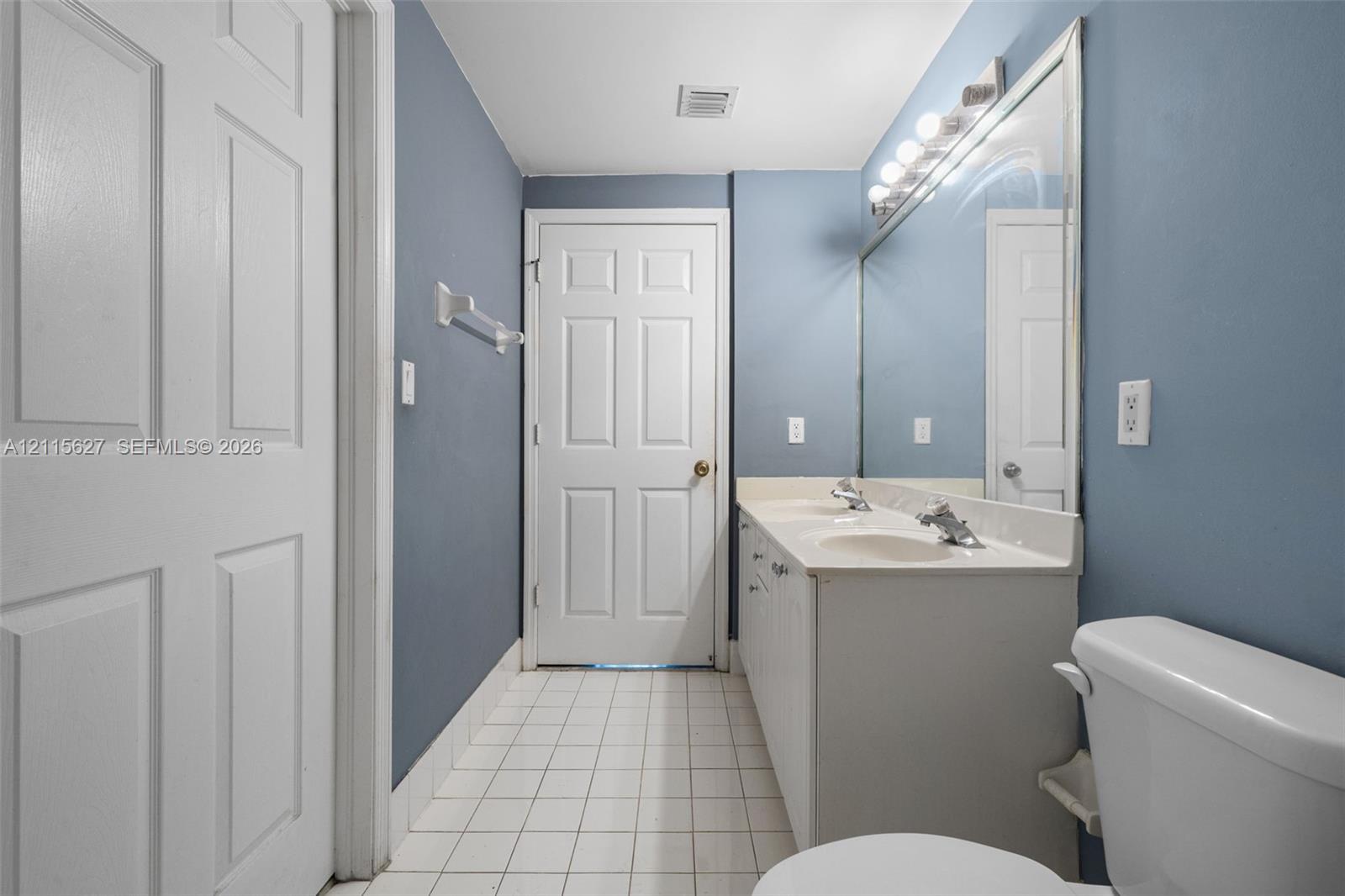 Dual-sink vanity with a solid-surface countertop, full-width vanity mirror, polished chrome fixtures, and tiled flooring