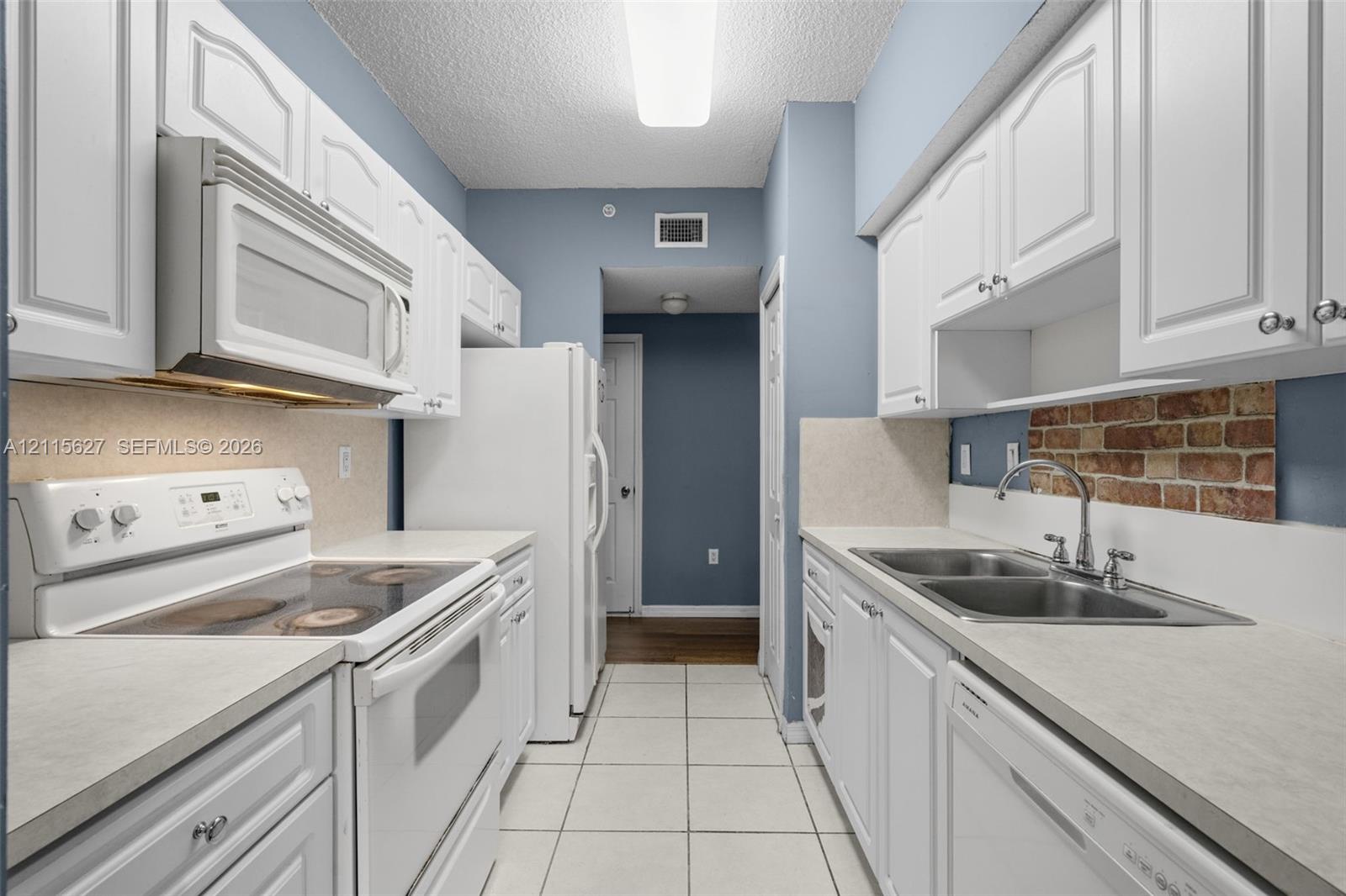 Galley kitchen featuring white cabinetry, light-toned countertops, tile flooring, an exposed brick backsplash, and a double-basin stainless steel sink