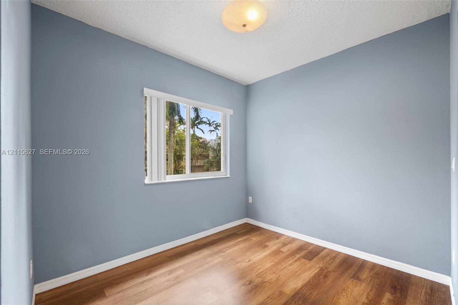 Room featuring wood-finish flooring, neutral blue-toned wall paint, a single window with vertical blinds, and a central ceiling-mounted light fixture