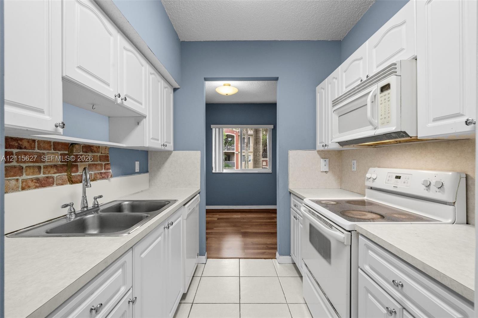 Kitchen featuring white shaker-style cabinetry, a brick backsplash, dual-basin stainless steel sink, tile flooring, and integrated white appliances
