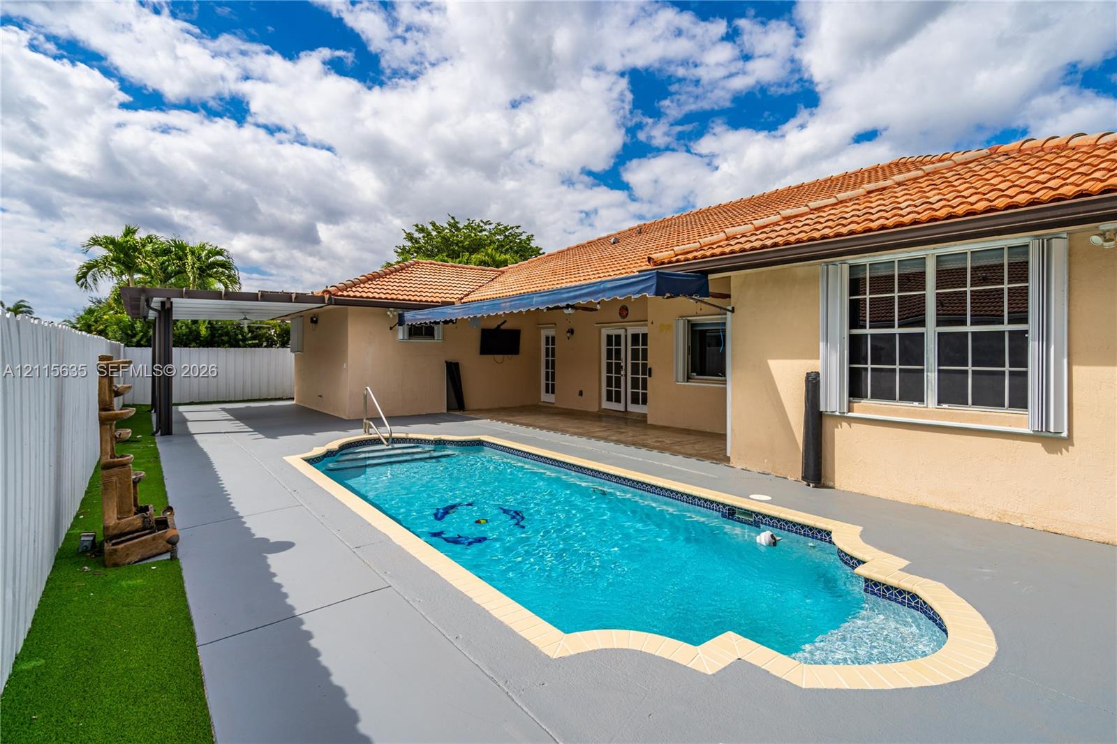 In-ground swimming pool with a custom-shaped perimeter, terra cotta tile roof, retractable fabric awning, wooden-frame pergola, and synthetic turf lawn
