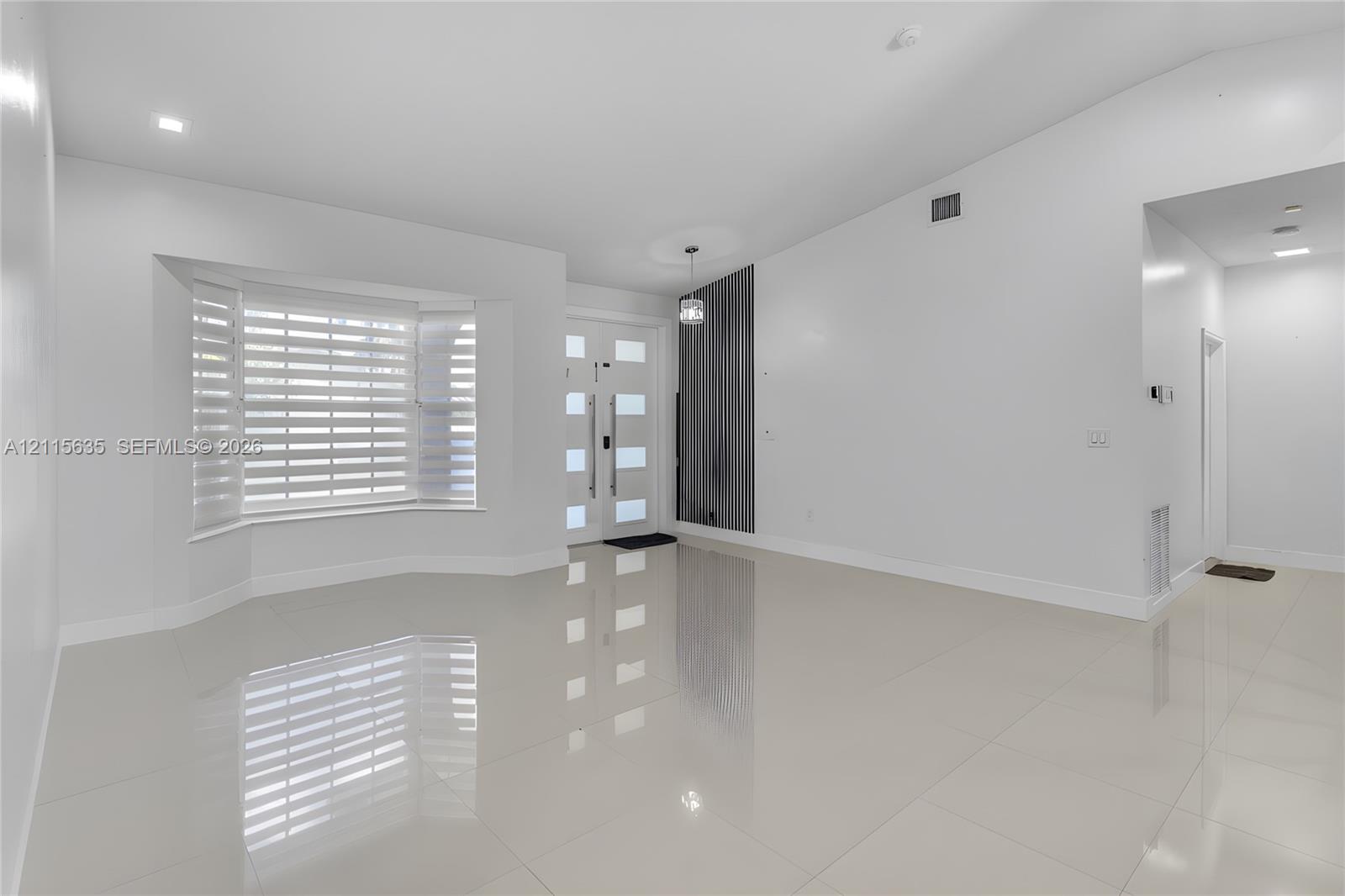 Bright interior featuring vaulted ceilings, polished tile flooring, a multi-pane front entry door with vertical handle hardware, and a bay window equipped with horizontal blinds