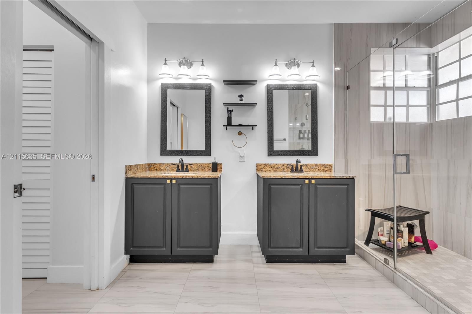 Dual-vanity bathroom featuring stone-surfaced countertops, charcoal-finish cabinetry, framed mirrors, and a glass-enclosed shower with tiled wall surrounds