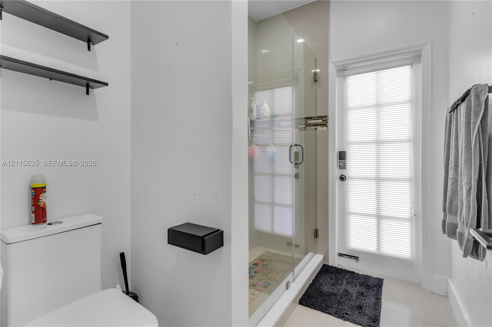 Full-height glass-enclosed shower, wall-mounted matte black shelving, matte black wall-mounted toilet paper holder, and a glass-paneled exterior door