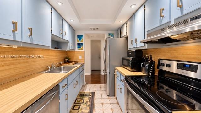 Galley kitchen featuring light blue cabinetry, gold-tone hardware, wood-finish countertops, stainless steel appliances, and recessed ceiling lighting