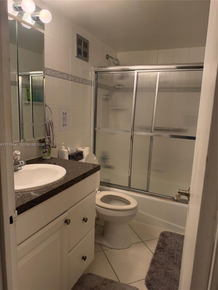 Bathroom featuring white cabinetry, dark stone-finish countertop with an integrated basin, tiled shower surround with sliding glass doors, and tiled flooring