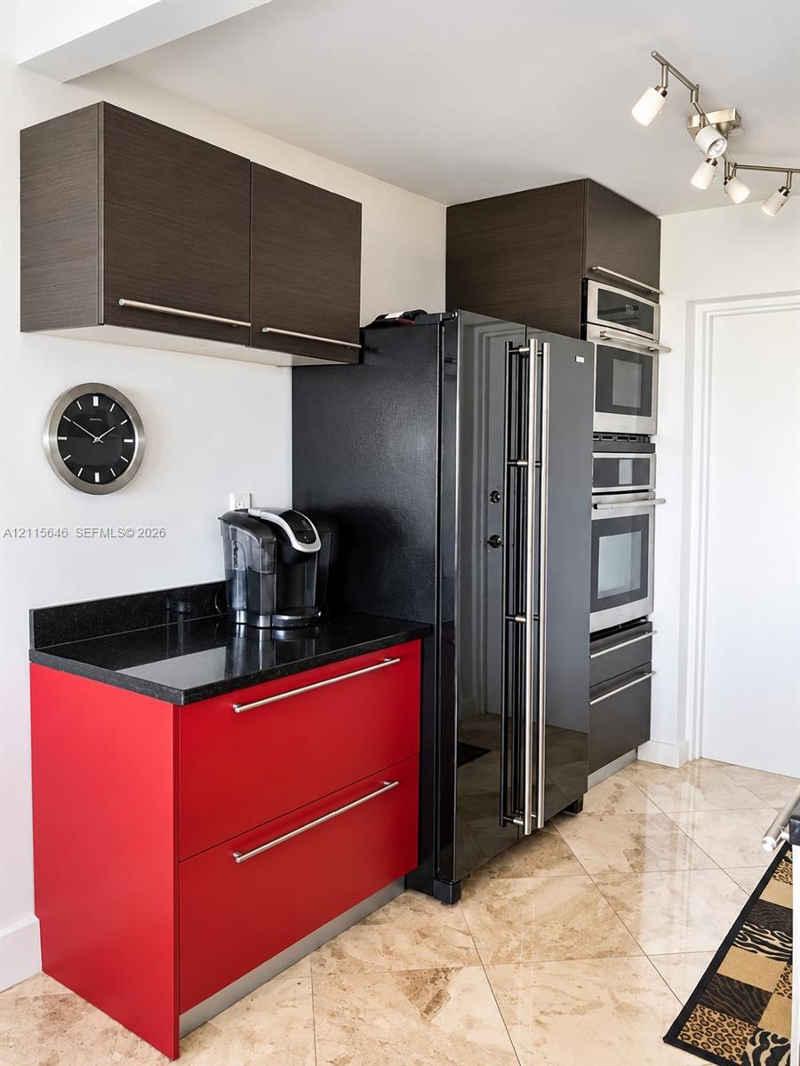 Kitchen area featuring flat-panel red lower cabinetry, dark wood-finish upper cabinetry, black countertops, stainless steel built-in double wall ovens, and tile flooring