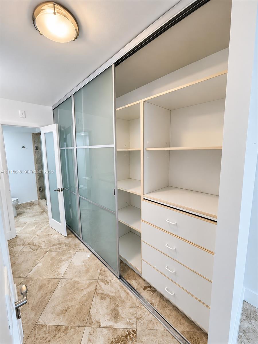 Custom closet featuring frosted glass sliding doors, built-in shelving, and a four-drawer dresser unit, accented by stone-finish floor tiling and flush-mount overhead lighting