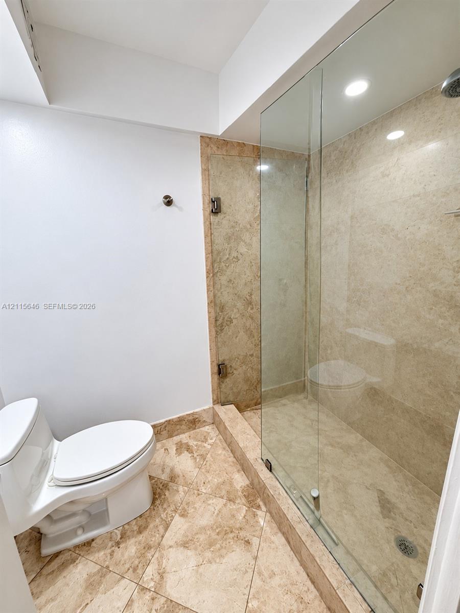 Walk-in shower with a glass enclosure, natural stone-finish wall tile, diagonal natural stone-finish floor tile, recessed lighting, and a dual-flush toilet