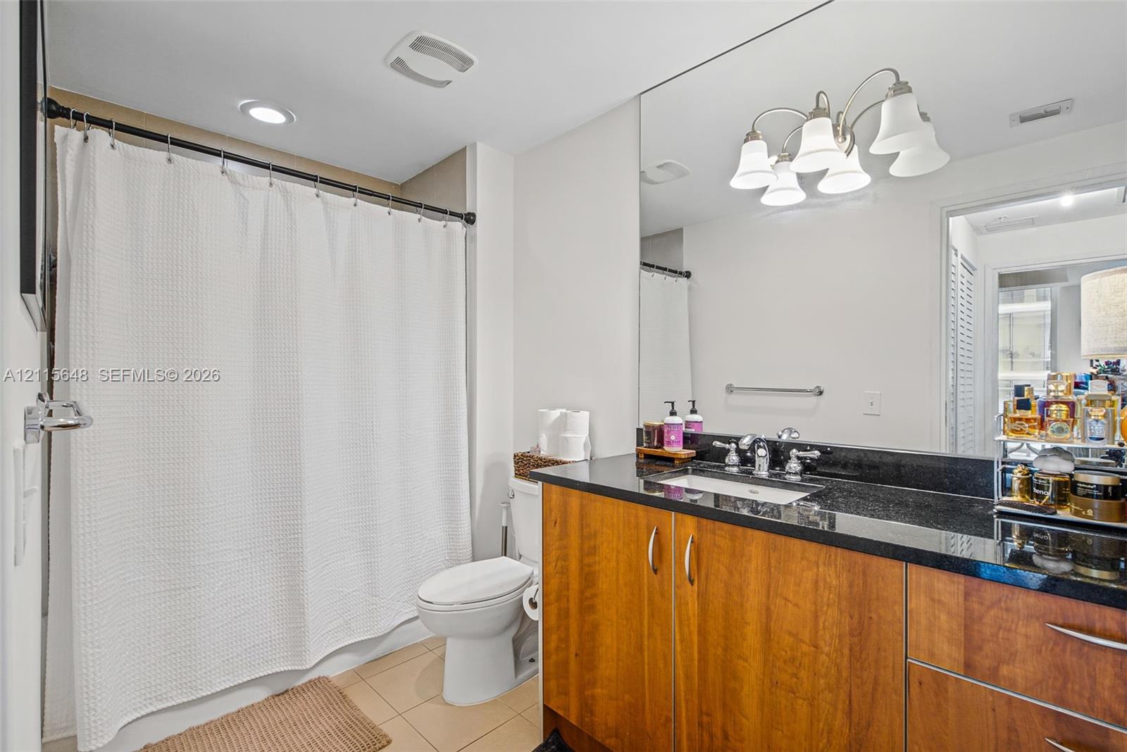 Bathroom featuring a dark-toned stone vanity countertop, wood-finish cabinetry, an undermount sink with dual-handle polished chrome faucet, a full-width wall mirror, and a five-light overhead fixture