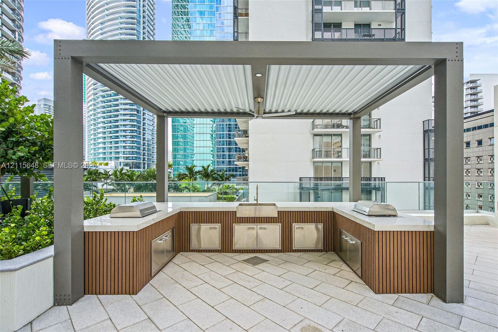 Outdoor kitchen featuring a structural metal pergola, stone-paver flooring, built-in stainless steel cabinetry, integrated cooktops, and a central basin