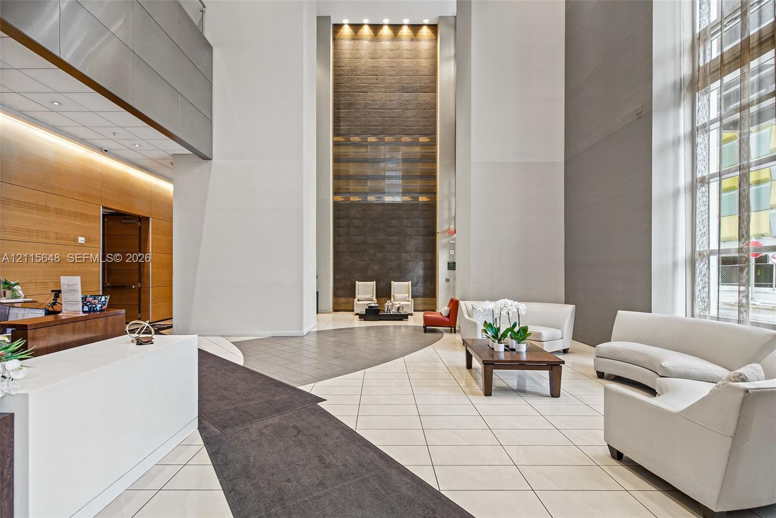Grand lobby featuring double-height ceilings, a floor-to-ceiling textured accent wall, polished stone flooring, recessed architectural lighting, and floor-to-ceiling windows