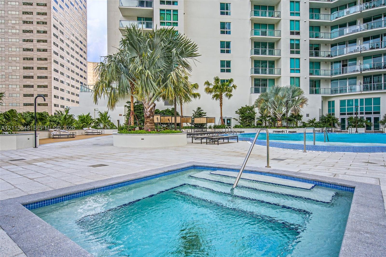 In-ground spa with stone-tile surround, expansive paved pool deck, large swimming pool, mature palm trees, and multi-story high-rise exterior