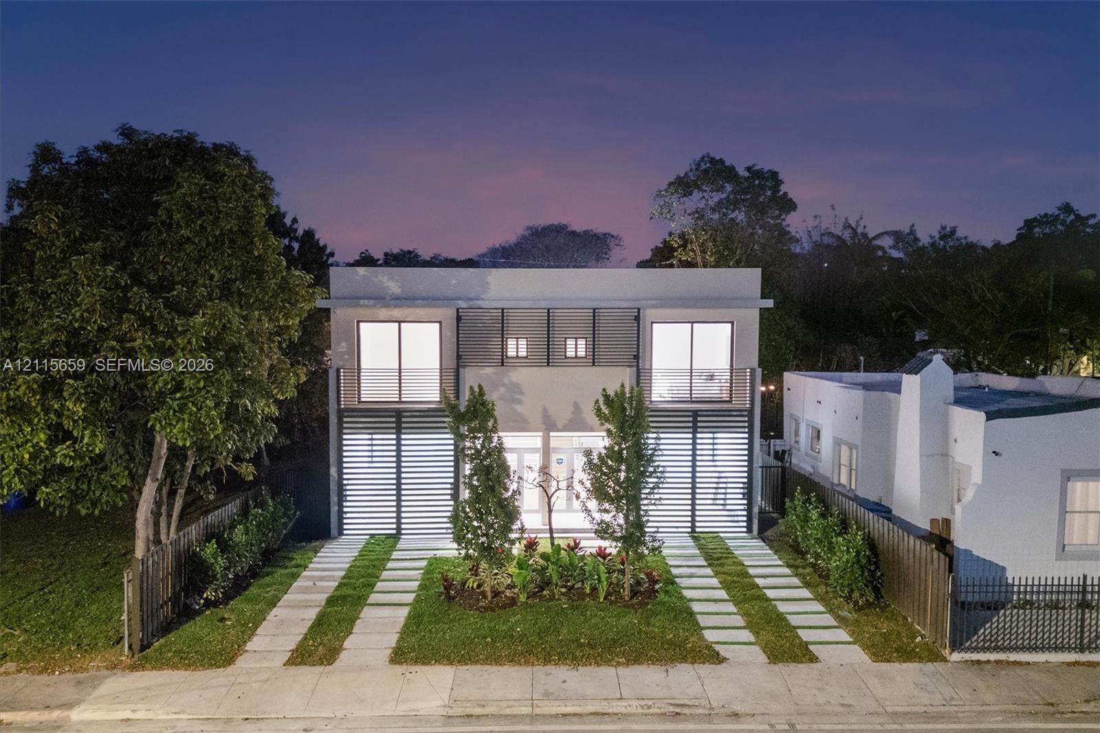 Modern two-story architectural exterior featuring horizontal slat privacy screens, large glass sliding doors, dual concrete-paver driveways, and native landscaping