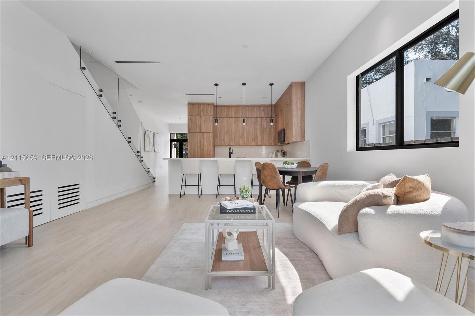 Open-concept interior featuring light wood-finish flooring, custom wood-veneer cabinetry, a center island with bar seating, and a glass-paneled staircase railing
