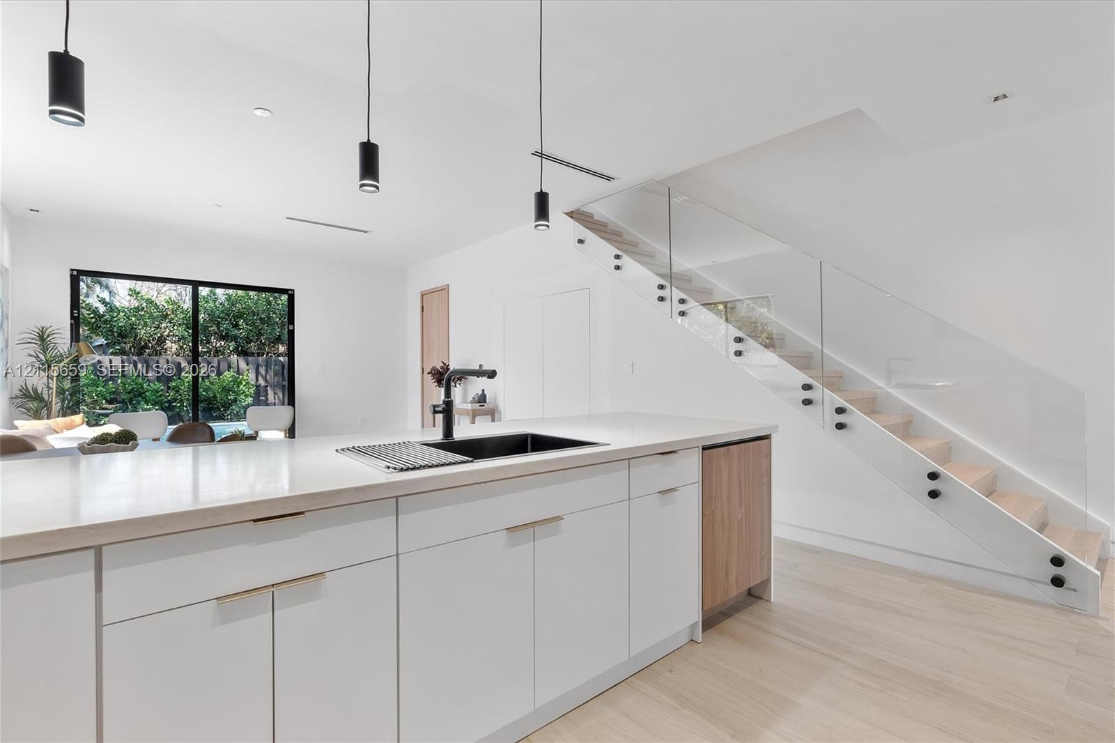 Modern kitchen island featuring a quartz countertop, black undermount sink, and matte black faucet, complemented by white cabinetry, wood-finish flooring, and a glass-paneled staircase