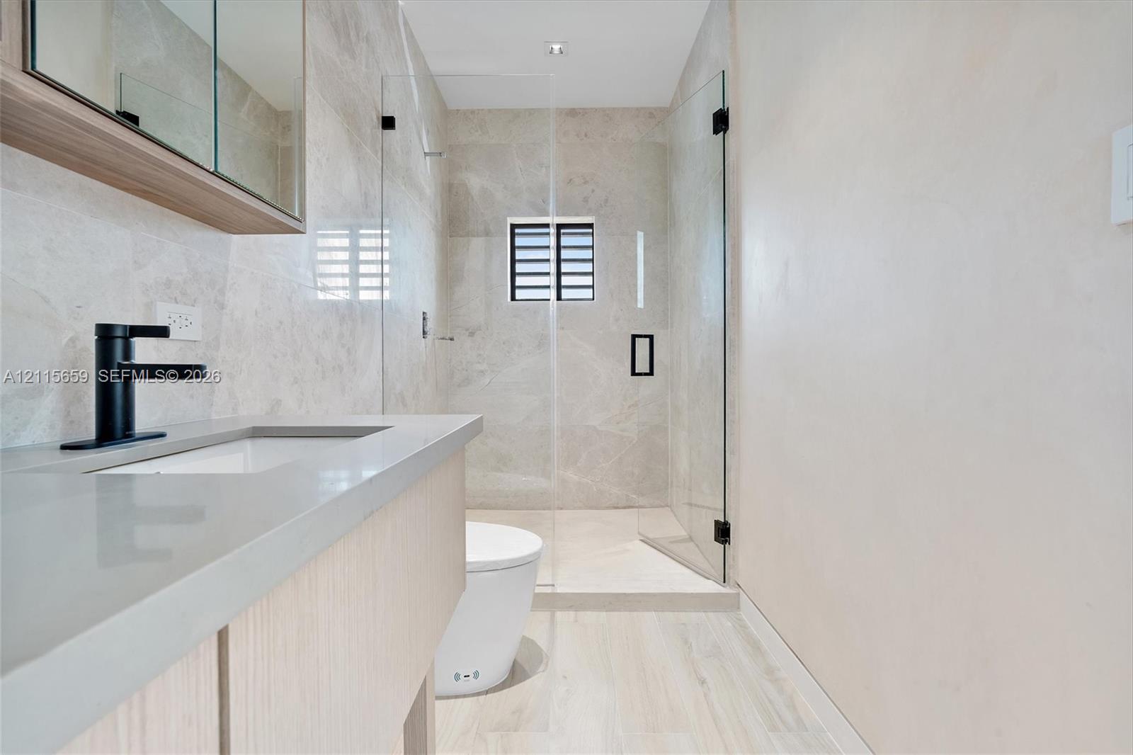 Bathroom featuring a wood-finish floating vanity, matte black faucet, full-height tile shower enclosure with a glass door, and large-format tile flooring